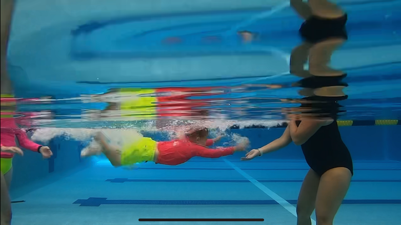 View from underwater of a boy in a pool swimming from one person to another. The boy is in the middle, facing the right, in neon yellow swim trunks and a neon red rash guard.